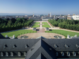 Über dem Dach des Karlsruher Schlosses fällt der Blick auf die Innenstadt von Karlsruhe. Im Vordergrund sind Park- und Waldanlagen zu erkennen sowie Teile des Schlossdaches.