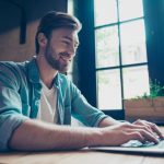 Side profile low angle shot of cheerful man smiling, searching information on his device in nice modern work station, in casual smart wear, grin, big windows with green nature view