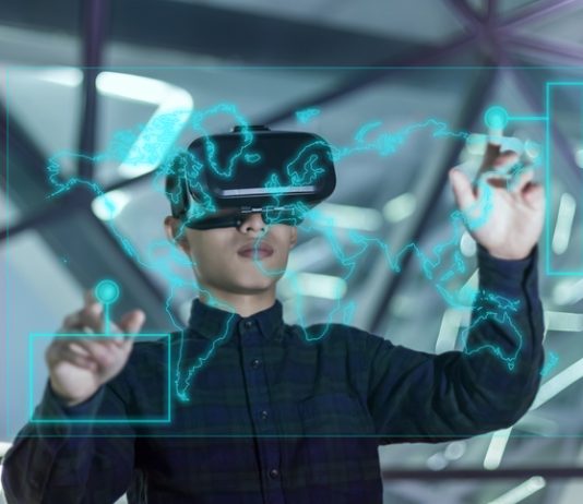 Mann mit VR Brille steht vor Glasscheibe, auf der mit grünen Linien eine Weltkarte gezeichnet ist. Der Mann berührt mit seinen Fingern die Karte. Sein Aussehen lässt darauf schließen, dass der Mann dem asiatischen Kulturkreis zuzurechnen ist.