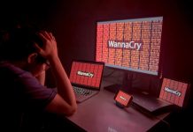 Mensch mit Brille, links im Anschnitt, stützt den Kopf in seine Hände: vor ihm vier verschiedene Monitore, unterschiedlicher Größe, auf denen zwischen einer roten 0-1-Zahlenfolge der Begriff WannaCry zu sehen ist.