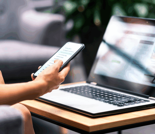 Frau mit Smartphone und modernem Notebook PC Computer auf dem Tisch schaut sich eine Website an.