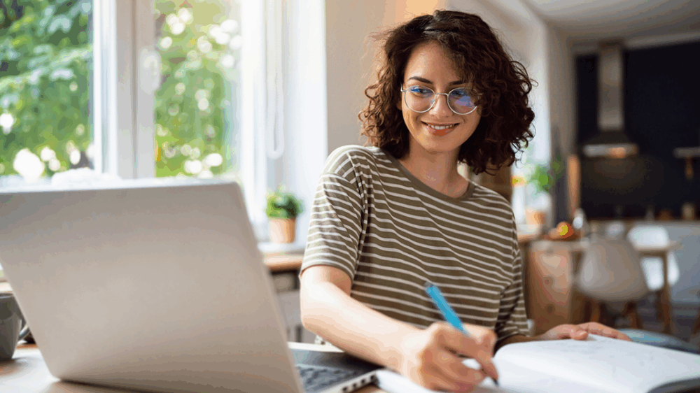 Junge Frau sitzt am Tisch mit Laptop und betreibt E-Learning.