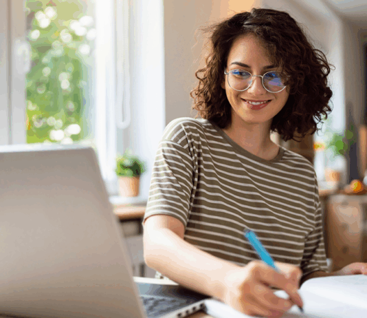 Junge Frau sitzt am Tisch mit Laptop und betreibt E-Learning.