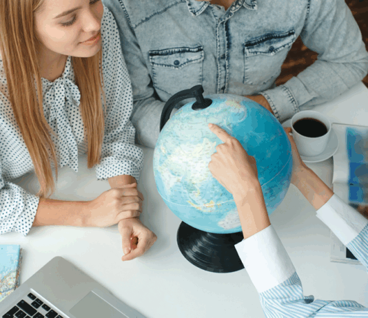 Junge Menschen in einem Reisebüro mit einem Reisebüro, das das Reiseziel auf einem Globus wählt und neugierig aussieht und lächelt.