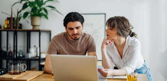 Symbolbild für Artikel: Warum der Weg zum Eigenheim so kompliziert geworden ist; Mann und Frau besprechen etwas und sitzen am Laptop